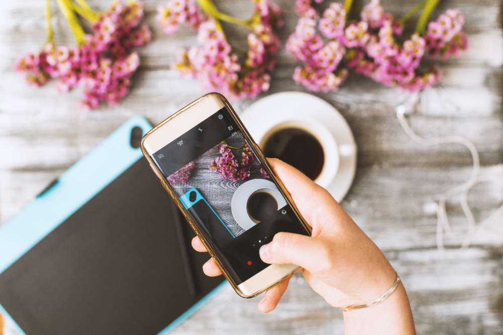 Instagram photographer blogging workshop concept, hand holding phone and taking photo of stylish flowers, cup of coffee and graphic tablet on grey wooden rustic background.space for text. Instagram photographer blogging workshop concept, hand holding phone and taking photo of stylish flowers, cup of coffee and graphic tablet on grey wooden rustic background.space for text